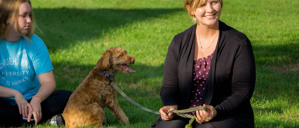 A woman sitting on the grass, smiling, while holding a book. Next to her, a girl in a blue T-shirt with 