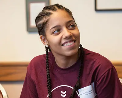 A person with braided hair smiles while looking to the side. They are wearing a maroon T-shirt and are sitting indoors.