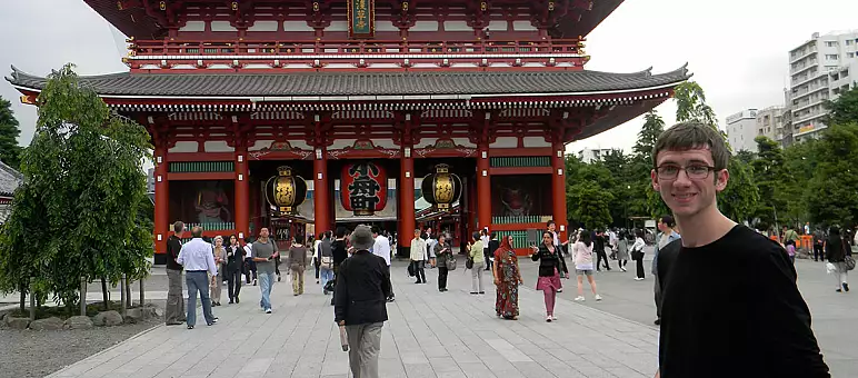 A person stands smiling in front of the Senso-ji Temple in Tokyo. The temple has a large red gate with lanterns, and many people are walking around in the courtyard. Trees and buildings are visible in the background.