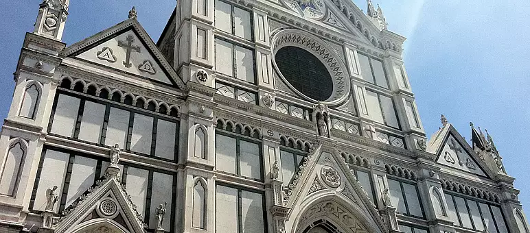 The image shows the facade of the Basilica of Santa Croce in Florence, Italy. It features Gothic architecture with ornate decorations, intricate carvings, and a large circular window at the center. The sky is clear and bright.