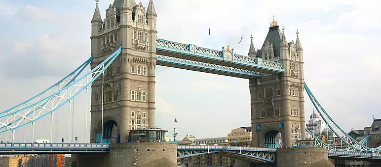 Tower Bridge in London on a cloudy day, featuring its iconic twin Gothic towers and blue suspension cables. The Thames River flows beneath the raised bascule. Buildings are visible in the background.