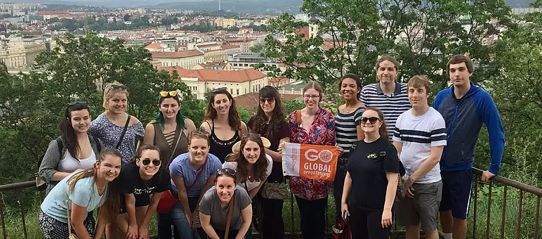 A group of people posing on a scenic overlook with a panoramic view of a city below. They are holding a 