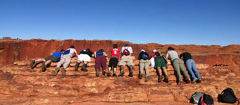 A group of people lying face down on a red rock formation under a clear blue sky. They are wearing casual clothes and backpacks. Some bags are placed on the ground nearby.