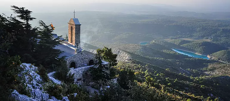 A stone building with a tower stands on a hill overlooking a vast landscape with rolling hills and a winding river below. The scene is dotted with greenery under a hazy sky, with sunlight casting soft shadows.