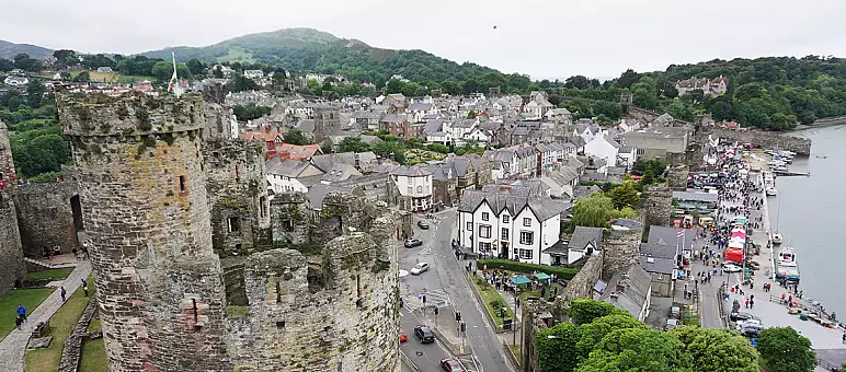 A scenic view of Conwy, Wales, featuring a medieval stone castle with circular towers. The town's quaint houses and narrow streets stretch along the waterfront. Green hills surround the area under a cloudy sky.