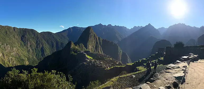 Panoramic view of Machu Picchu bathed in sunlight, with ancient stone structures and terraces nestled among lush green mountains. The sky is clear with a bright sun shining, casting shadows over the landscape.