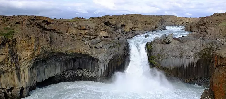 Panoramic view of a waterfall cascading between steep rocky cliffs, surrounded by textured basalt columns and rough terrain. The water below is turbulent, and the sky is cloudy, adding a dramatic effect to the scene.