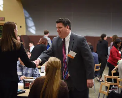 A man in a suit and colorful tie shakes hands with a woman at a networking event. People are seated at tables in the background, engaging in conversation. The setting appears to be a professional or conference environment.