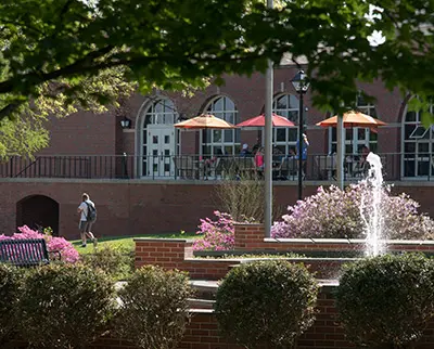 A scenic view of a brick building with large windows and a patio featuring colorful umbrellas. A fountain and flowering bushes are in the foreground. A person walks on a path, and trees frame the scene, casting shade.