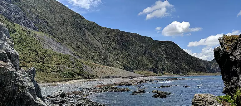 A scenic coastal view with rocky shores and crystal clear water under a bright blue sky. Steep, green hills rise from the shoreline on the left, while fluffy white clouds dot the sky. The sea is calm with scattered rocks near the shore.