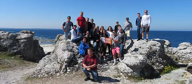 A group of people posing on large rocks with the sea in the background. The sky is clear and blue. One person holds an orange sign that reads 