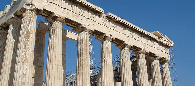 View of the Parthenon in Athens, Greece, featuring its iconic ancient columns and classical architecture. Scaffolding is visible around the structure, set against a clear blue sky.