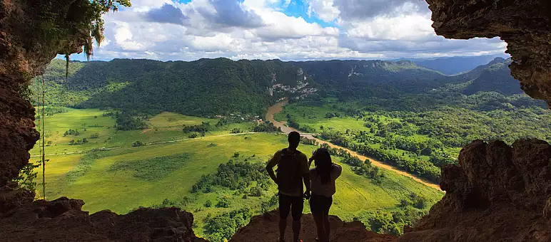 Two people stand at the entrance of a cave, overlooking a scenic view of a lush green valley with a winding river. The sky is partly cloudy, providing a dramatic backdrop to the expansive landscape.