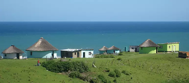 A scenic view of traditional round huts with thatched roofs and a few rectangular houses on grassy hills, overlooking a vast, calm ocean under a clear blue sky.