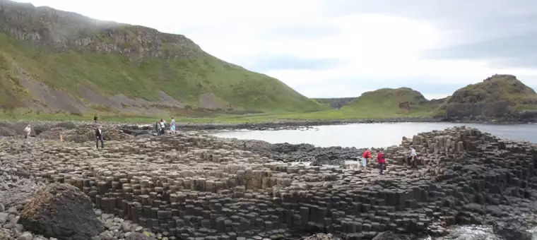 People exploring the Giant's Causeway in Northern Ireland, featuring interlocking basalt columns on a rugged coastline. Green hills rise in the background under a cloudy sky.