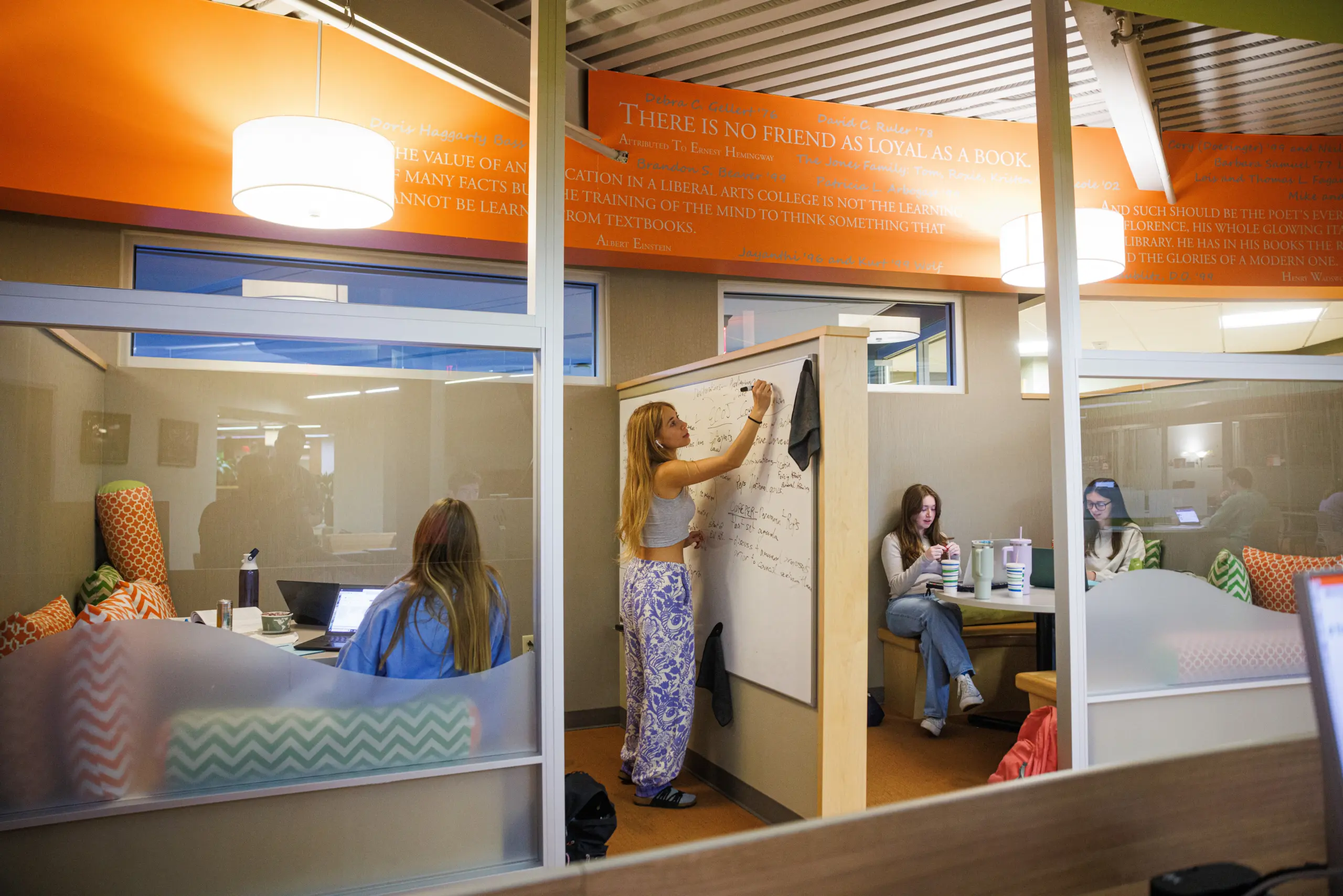 A student writes on a whiteboard inside a glass-walled study area. Other students sit around on couches with laptops and notebooks. The room has orange accents and inspirational quotes on the walls.