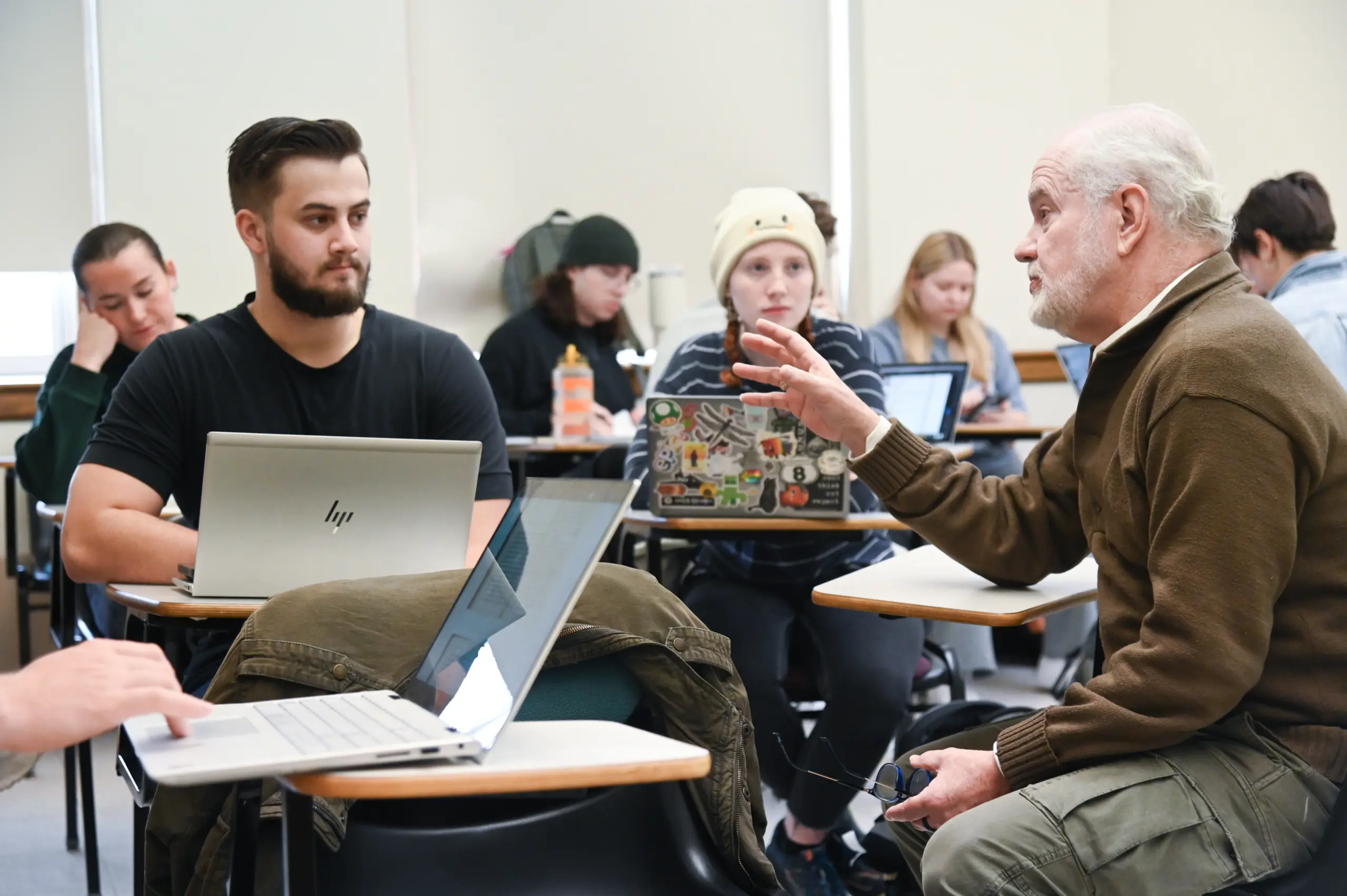 A classroom scene with a bearded older man talking to a younger man seated at a desk with a laptop open. Other students in the background are focused on their laptops and notebooks. The room has bright lighting and neutral-colored walls.
