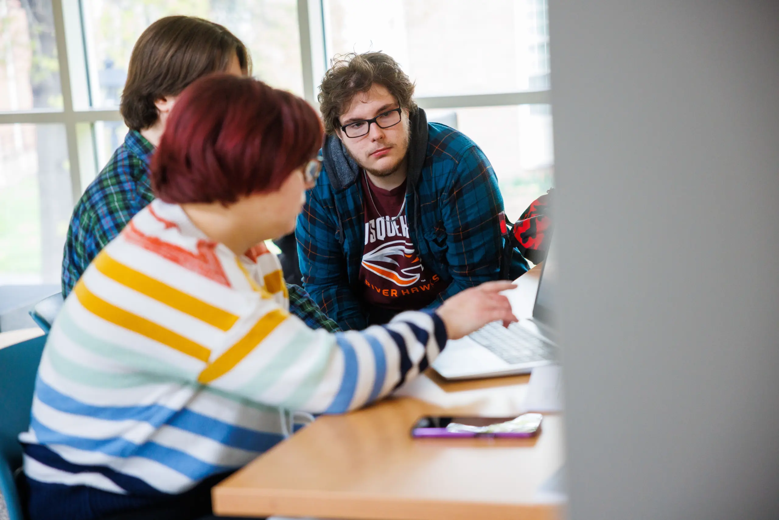 Three people are gathered around a table, looking focused on a laptop. One person with red hair is pointing at the screen while others watch attentively. The setting appears to be a bright room with large windows.