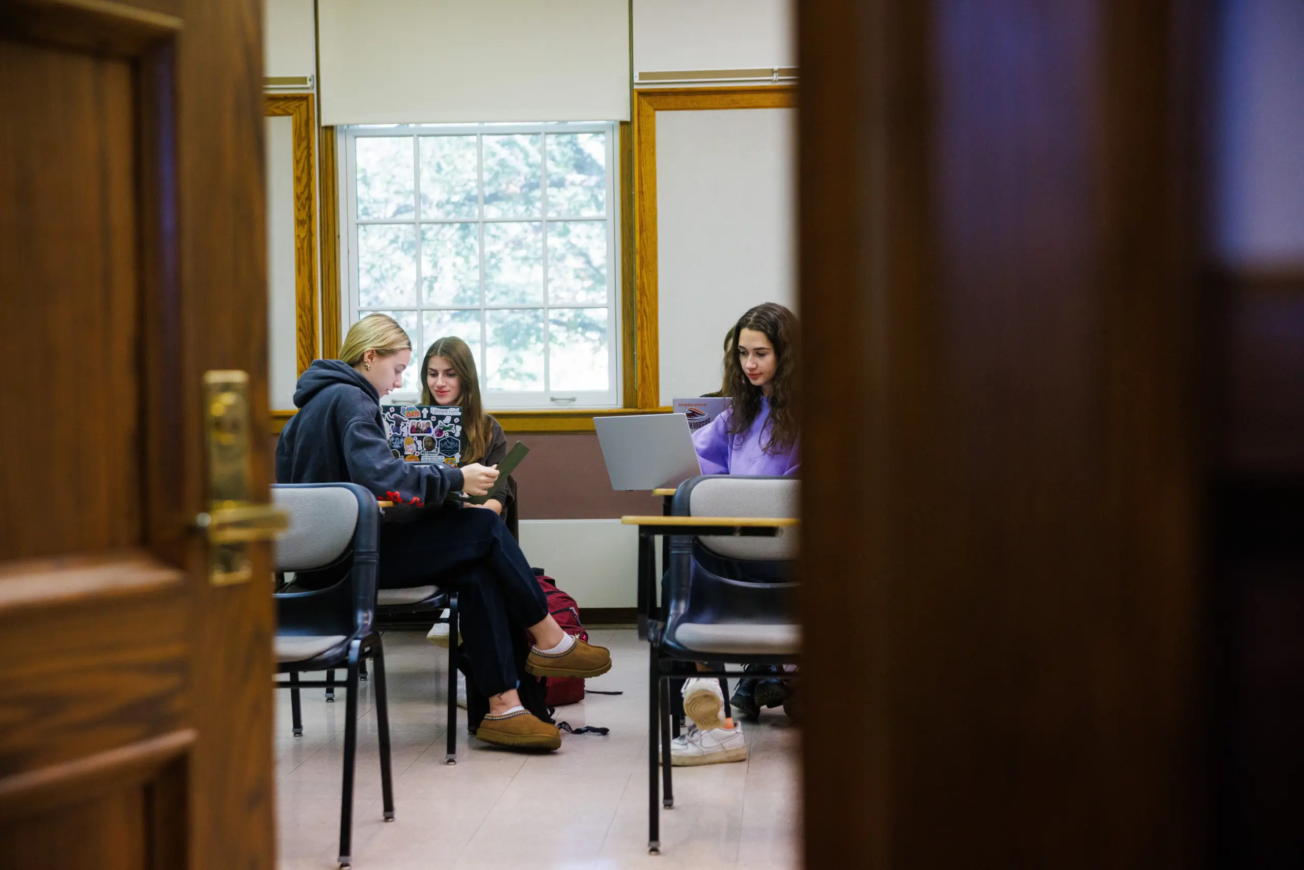 Three people are seated in a classroom setting. They are engaged with laptops and notebooks. The scene is viewed through an open door, with a window in the background allowing natural light in.