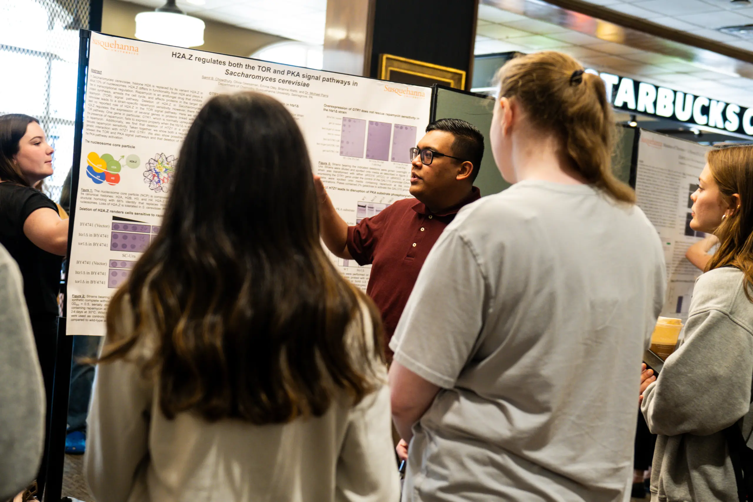 A group of people stand indoors, engaged in listening to a man in a maroon shirt presenting a research poster. The setting appears to be a conference or academic event, with a Starbucks visible in the background.