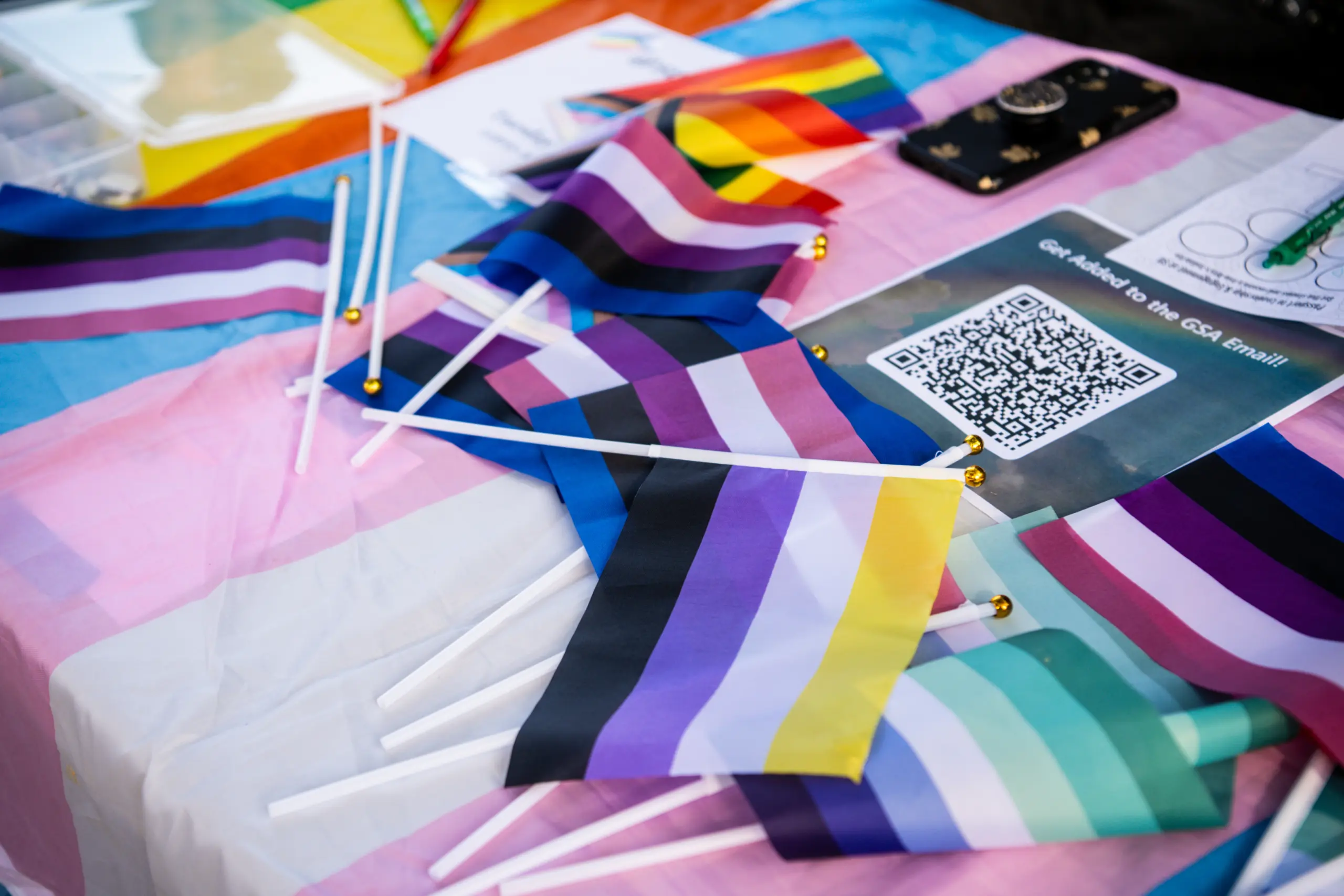 A table is covered with various pride flags, including the transgender and non-binary flags. A QR code and phone on the table are visible. The background has colorful materials suggesting a festive or activist setting.