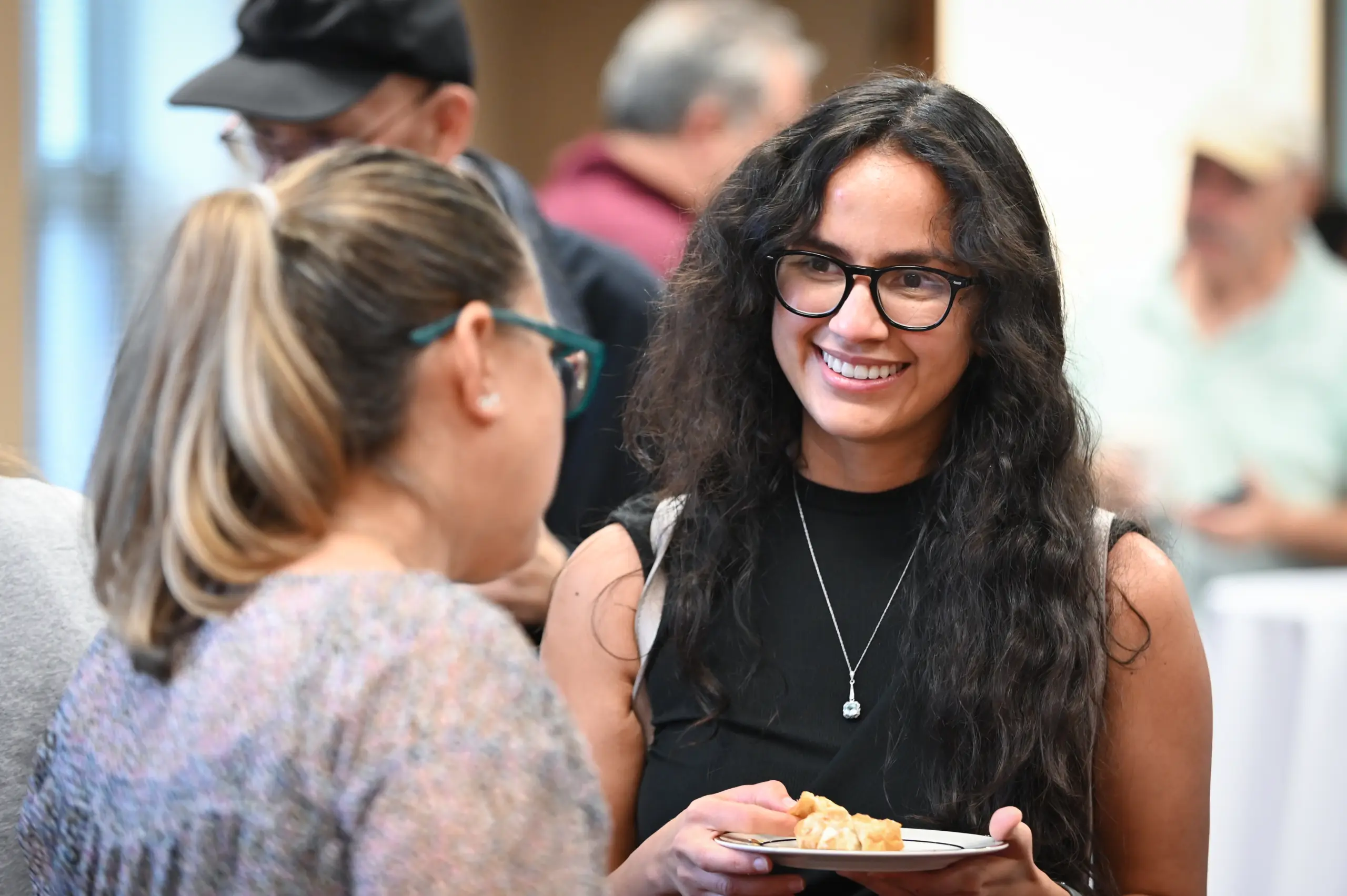 A woman with long curly hair and glasses smiles while holding a plate of food. She is talking to another woman with her hair in a ponytail. The background shows other people in a casual indoor setting.