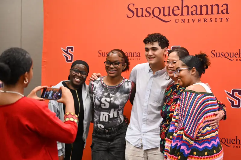 A group of five people are posing and smiling for a photo in front of an orange 体育买球 University backdrop. A person in a red outfit is taking their picture with a smartphone.