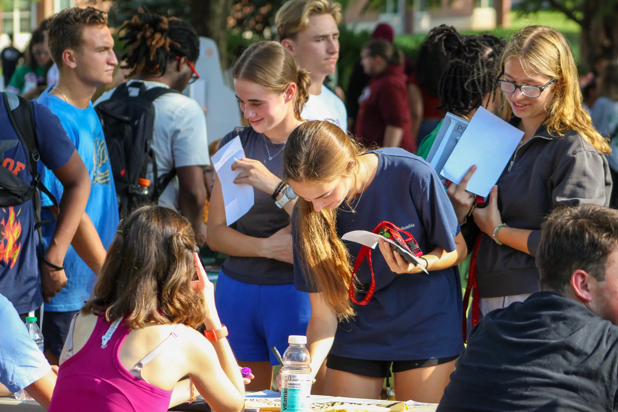 A crowd of students gathers around a table at an outdoor event. Some hold notebooks and chat, while one person leans over to sign something. Backpacks are visible, and sunlight casts shadows on the scene, creating a lively, engaging atmosphere.