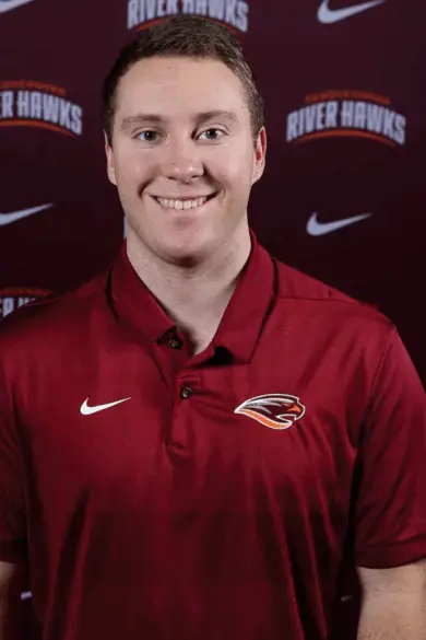 A person in a maroon polo shirt with embroidered logos stands smiling in front of a maroon backdrop with 