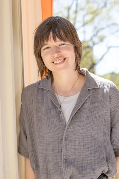 A person with short, brown hair smiles while leaning against a pillar. They wear a gray, short-sleeved shirt over a white top. The background features trees and a blurred outdoor setting with bright sunlight.