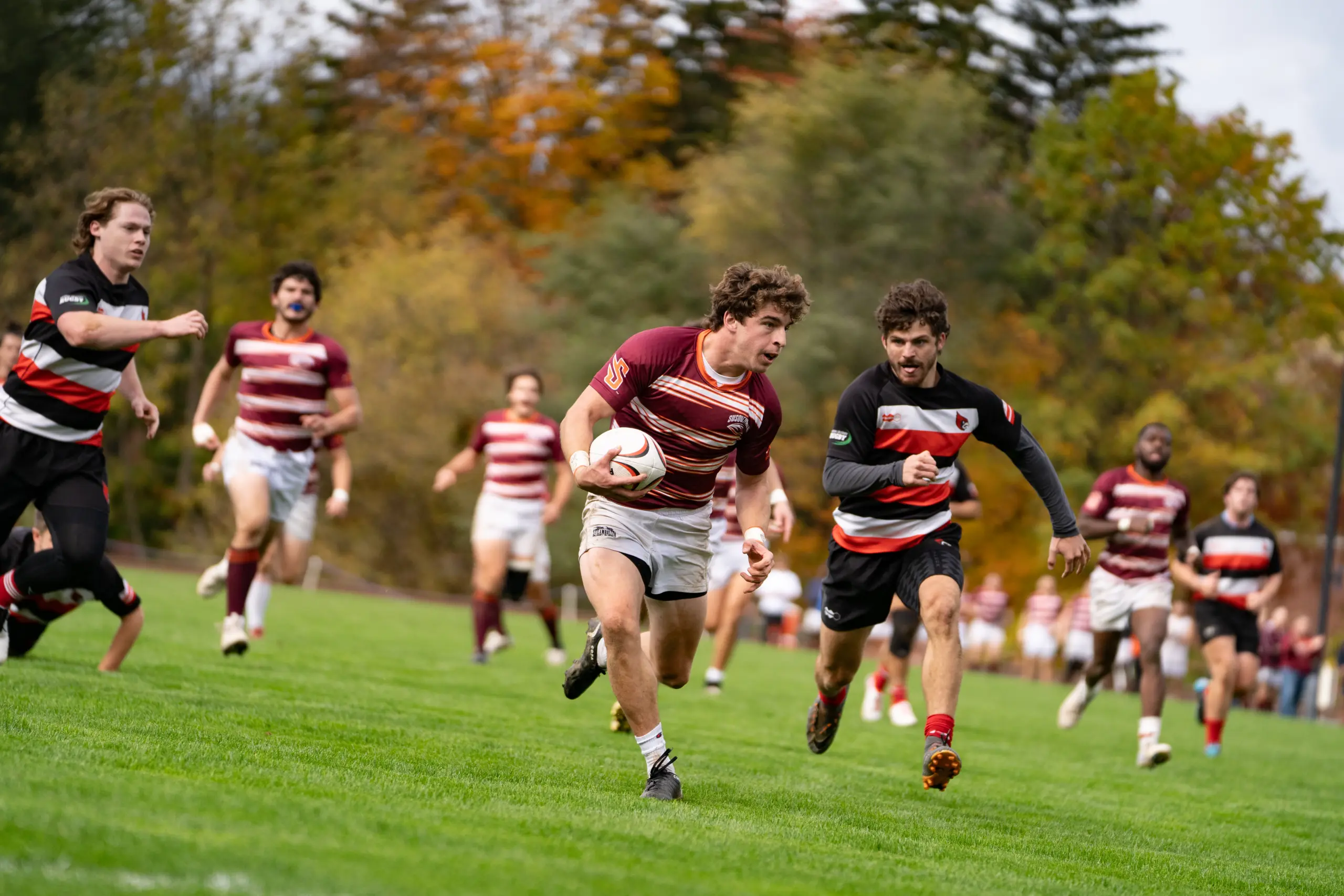 A male rugby player in maroon and white, embodying the spirit of athletics, runs with a ball on a grassy field, pursued by an opponent in black and red. Other players in team uniforms are visible against a backdrop of trees and autumn foliage, capturing the essence of outdoor recreation.