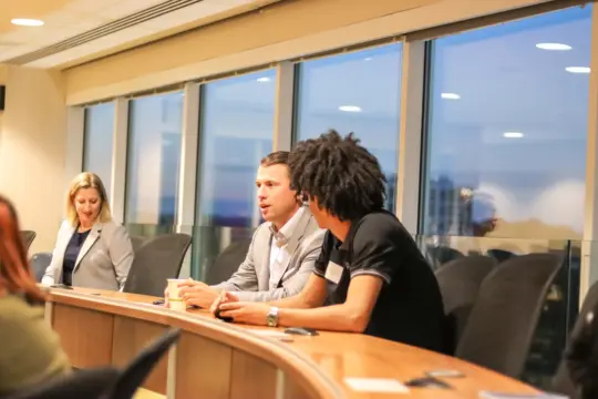 Three people seated at a long desk in a modern office setting, engaged in conversation. One person holds a cup, while others are listening and chatting. Large windows reveal an evening cityscape outside.