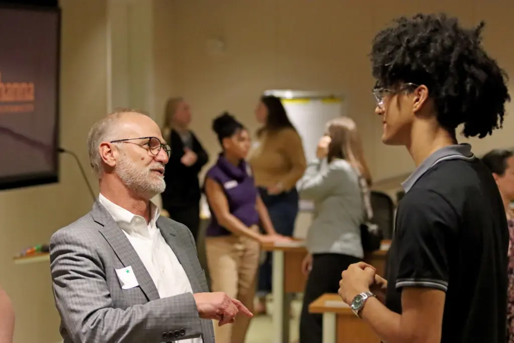 Two individuals are engaged in conversation in an indoor setting. The older man wears a gray suit, and the younger man has curly hair and glasses. A group of people are socializing in the blurred background.