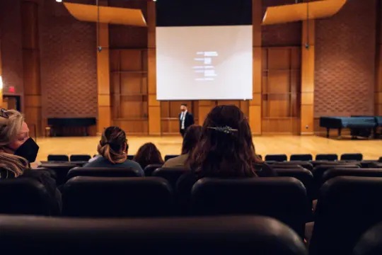 Audience members seated in a theater, facing a person standing by a screen displaying text. The room has brick walls and a wooden stage with a grand piano to the side.