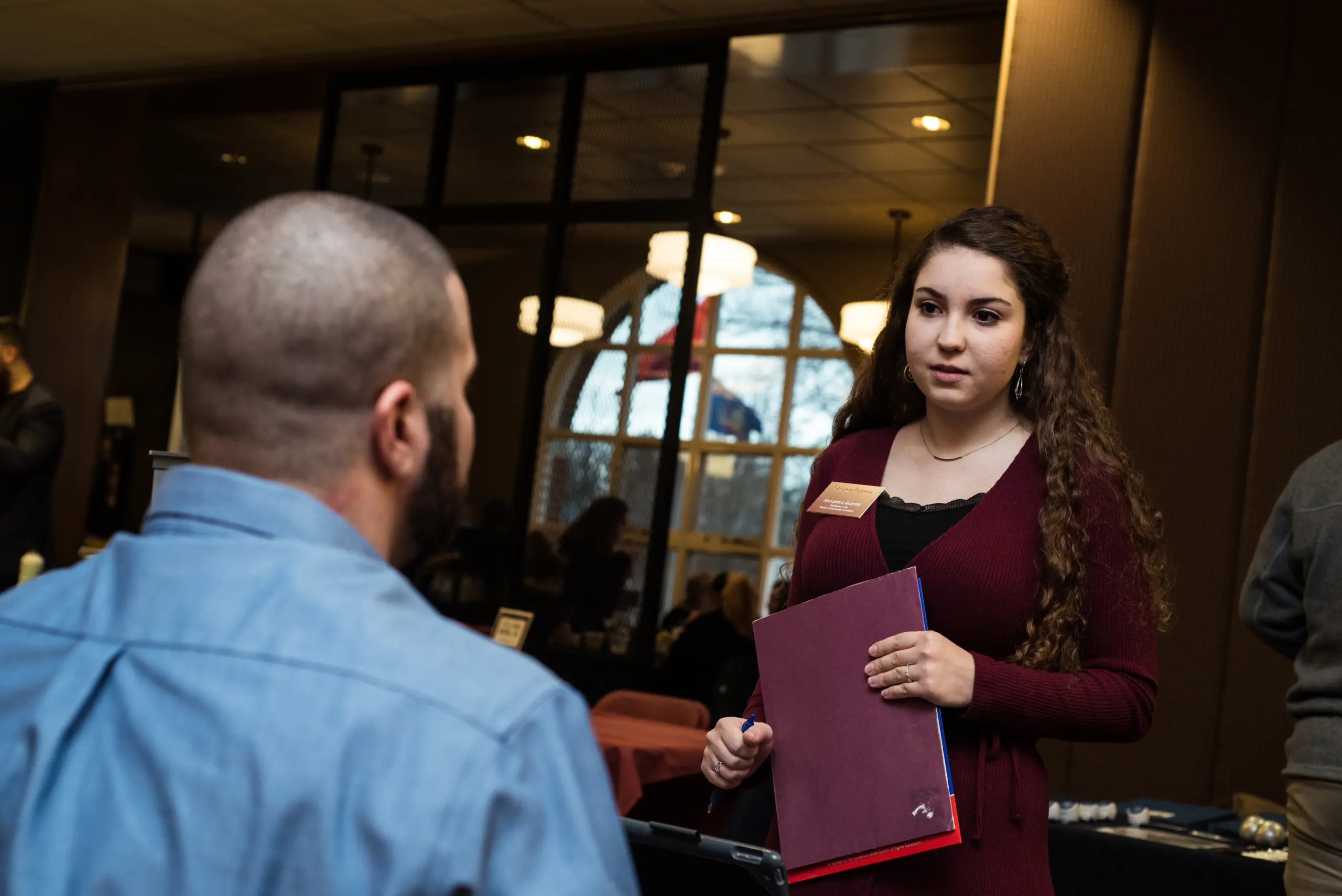 A woman with long curly hair, wearing a maroon cardigan, holds a portfolio and stands in front of a man in a blue shirt, engaged in conversation. They are in a room with large windows and multiple tables.