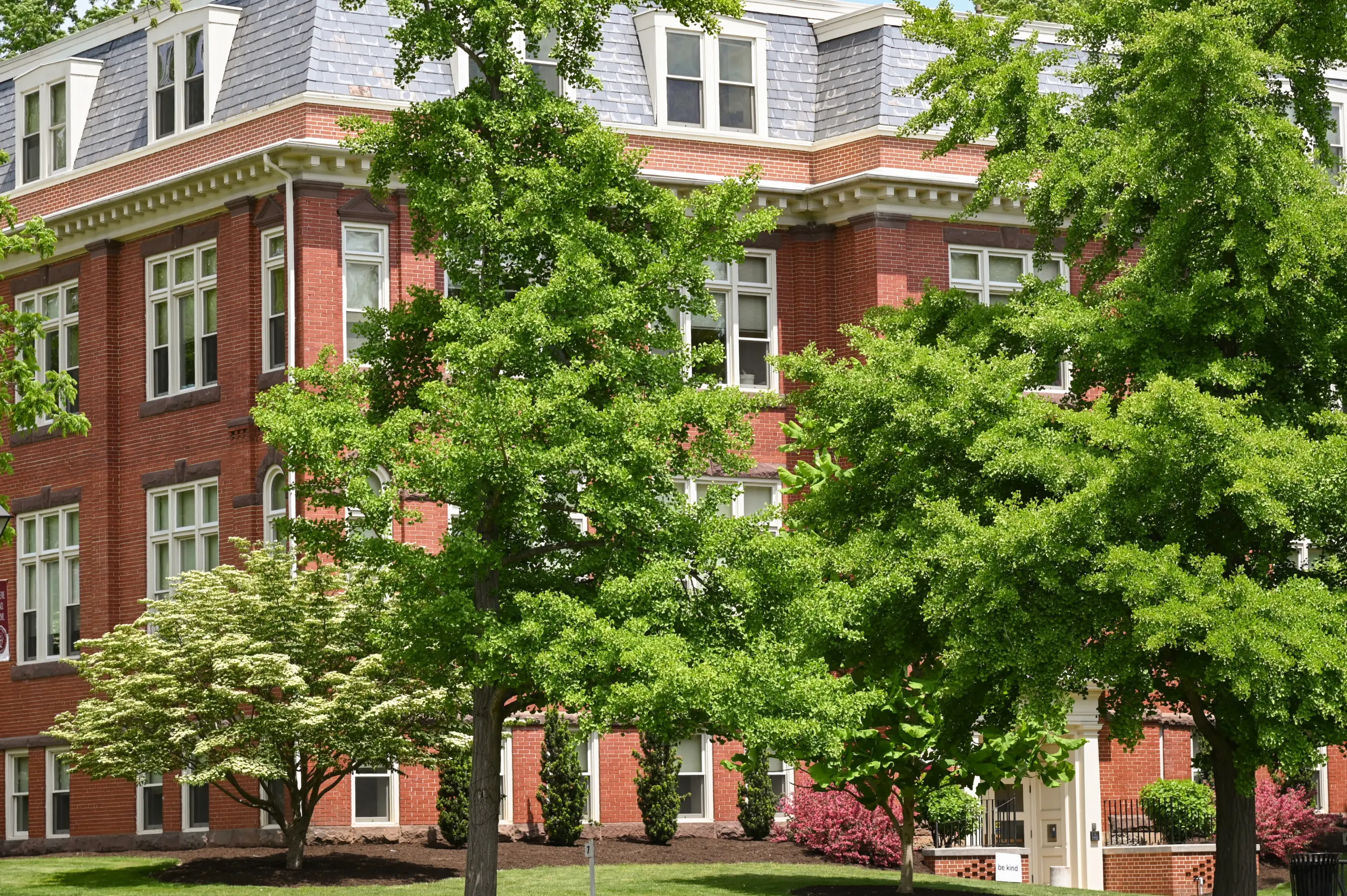 A large red brick building with white trim, partially obscured by lush green trees and shrubs. The building has numerous tall windows and a slate roof. The scene is bright and sunny, with well-maintained grass and landscaping.