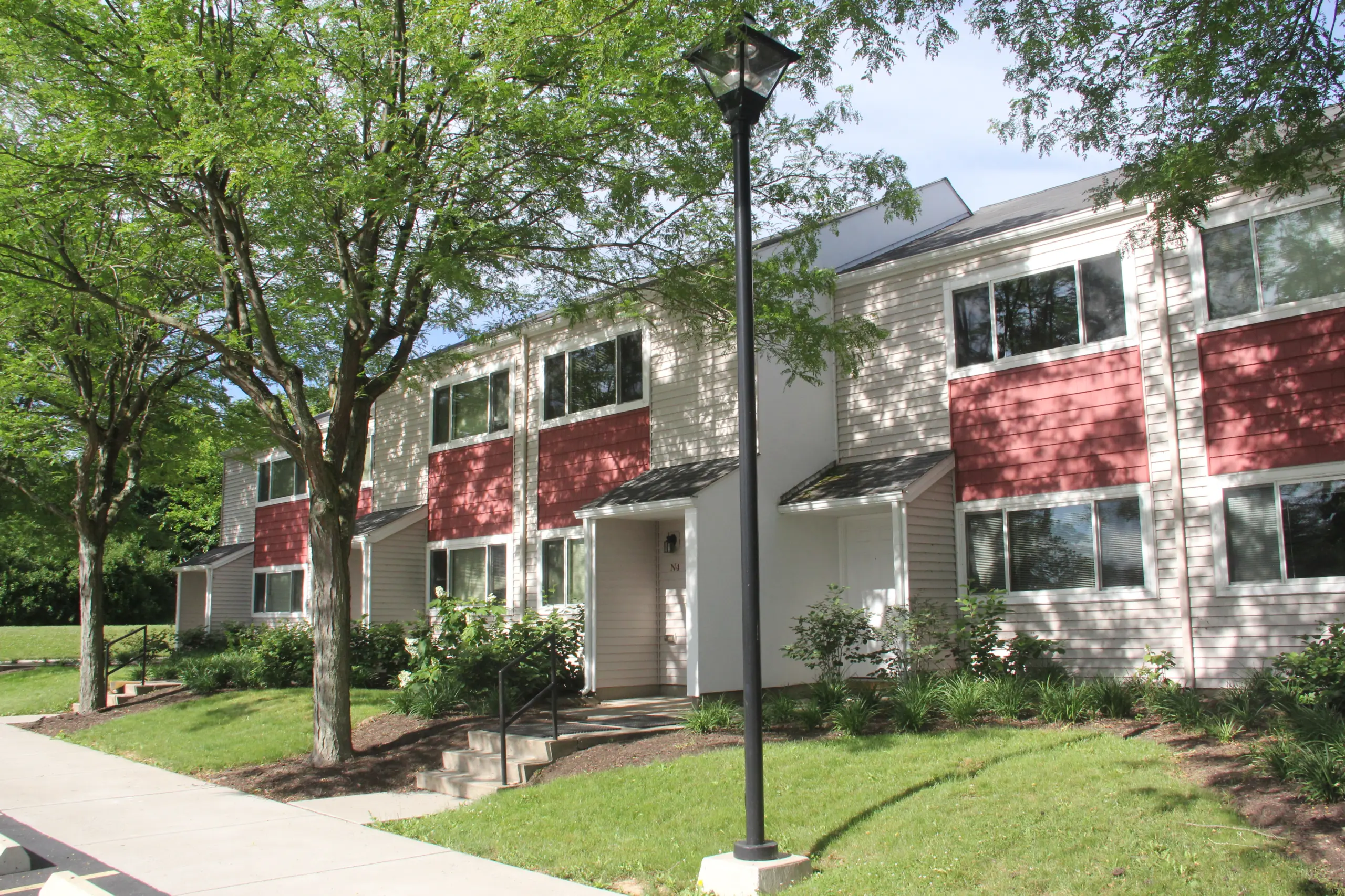 A row of modern townhouses with red and white exteriors, bordered by green lawns and trees. A lamppost stands on a paved walkway in the foreground. The area appears sunny and well-maintained.