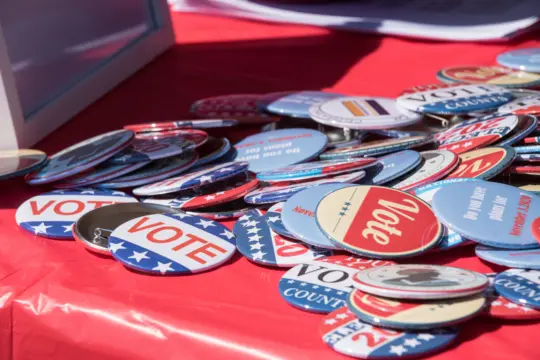 A collection of round, colorful buttons scattered on a red tablecloth. The buttons feature words like 