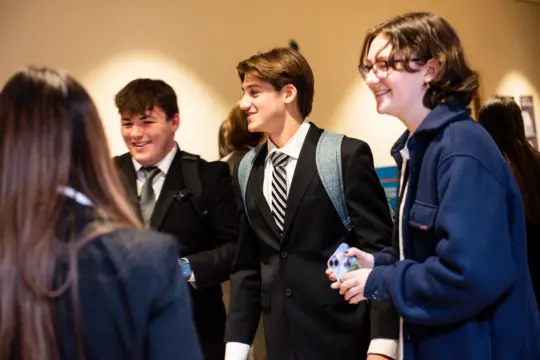 A group of four young people smiling and talking. Two of them are wearing suits, one has a backpack, and another is holding a smartphone. They appear to be in a warmly lit indoor setting.