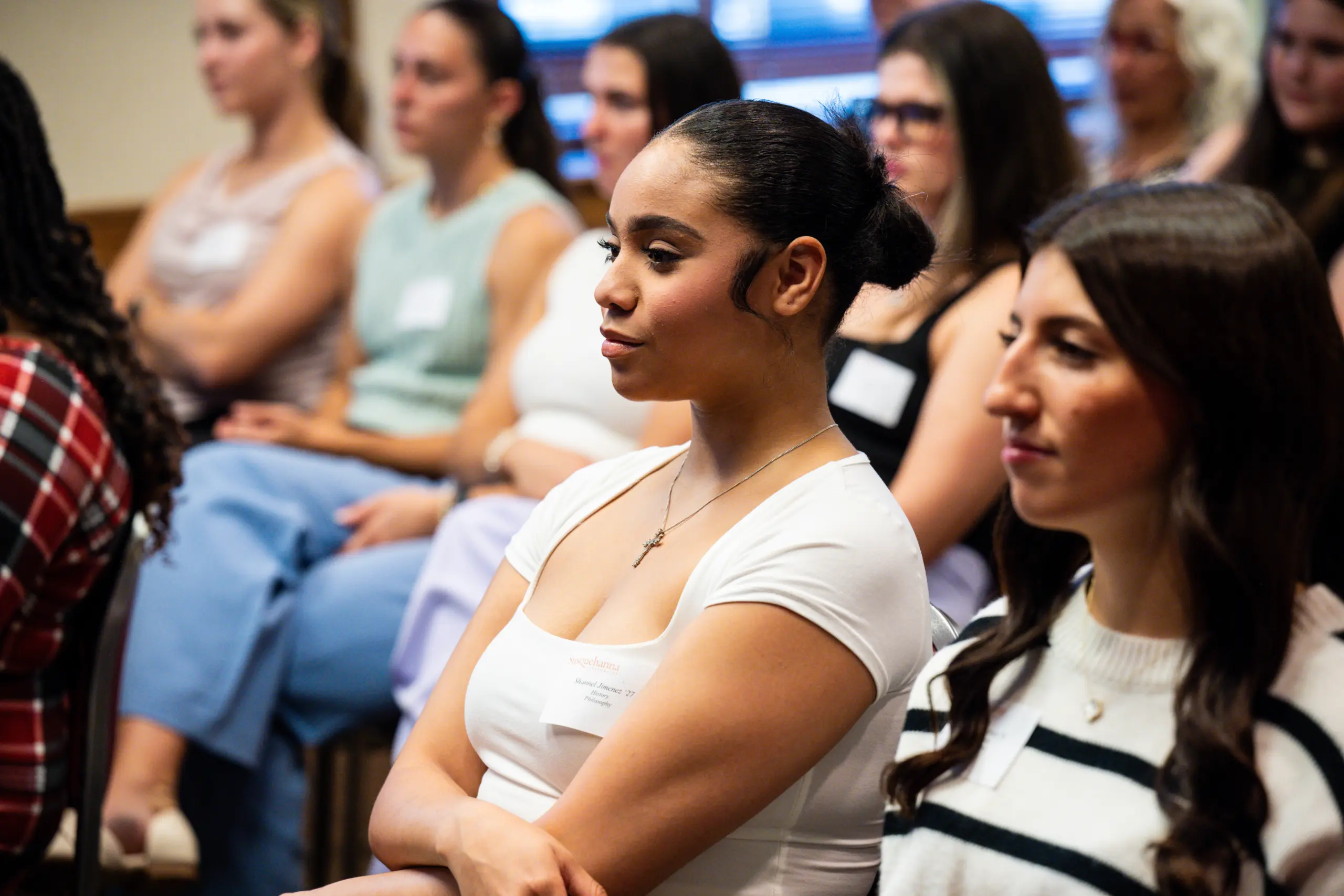 A group of people seated in a conference room, attentively listening to a speaker. The focus is on a person with dark hair tied in a bun and wearing a white shirt. Another person in the foreground has long dark hair and a striped sweater.