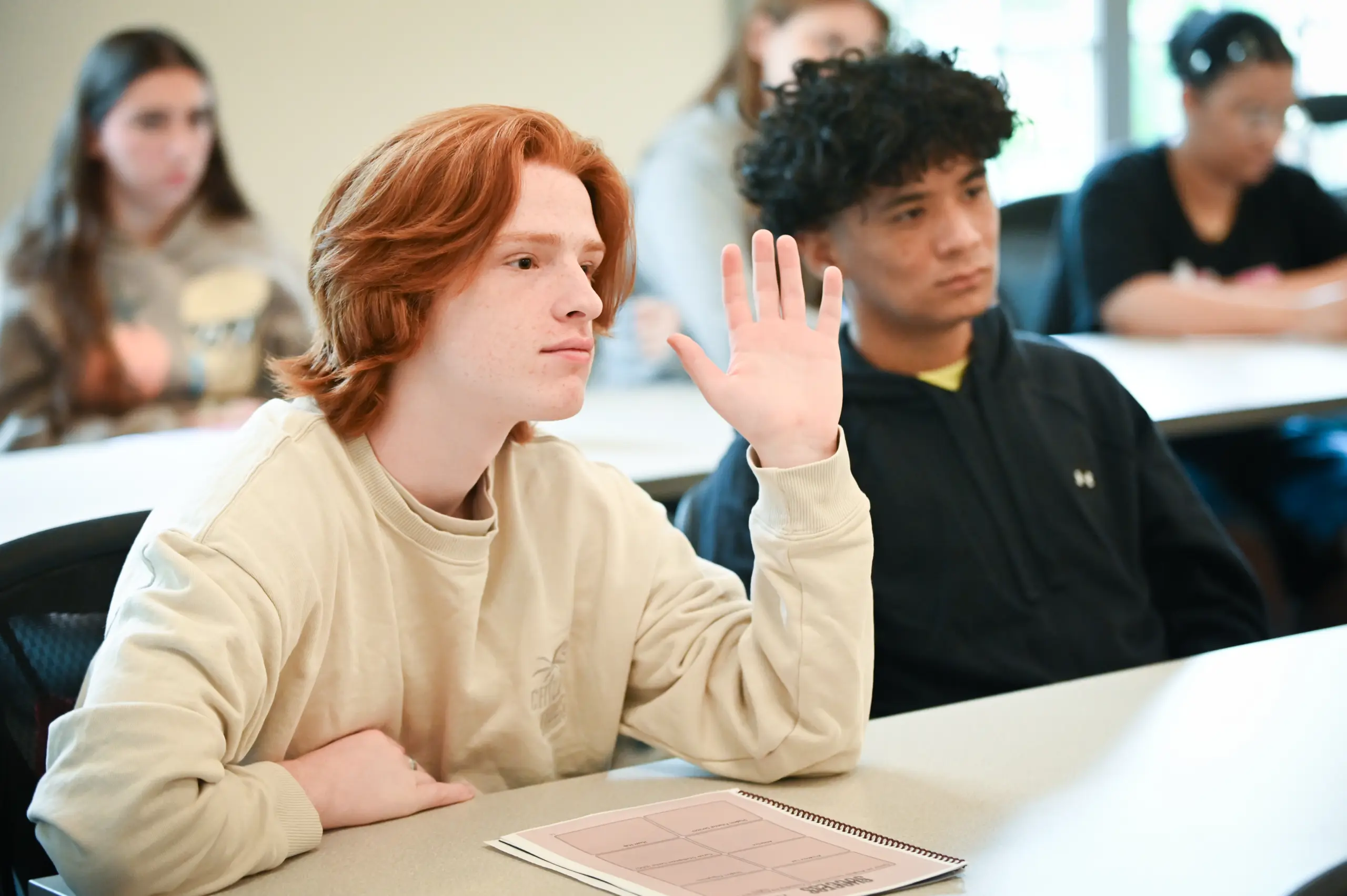 A young person with red hair raises their hand in a classroom, sitting next to another student with dark curly hair. Both are at desks, with notebooks in front of them. More students sit in the background.