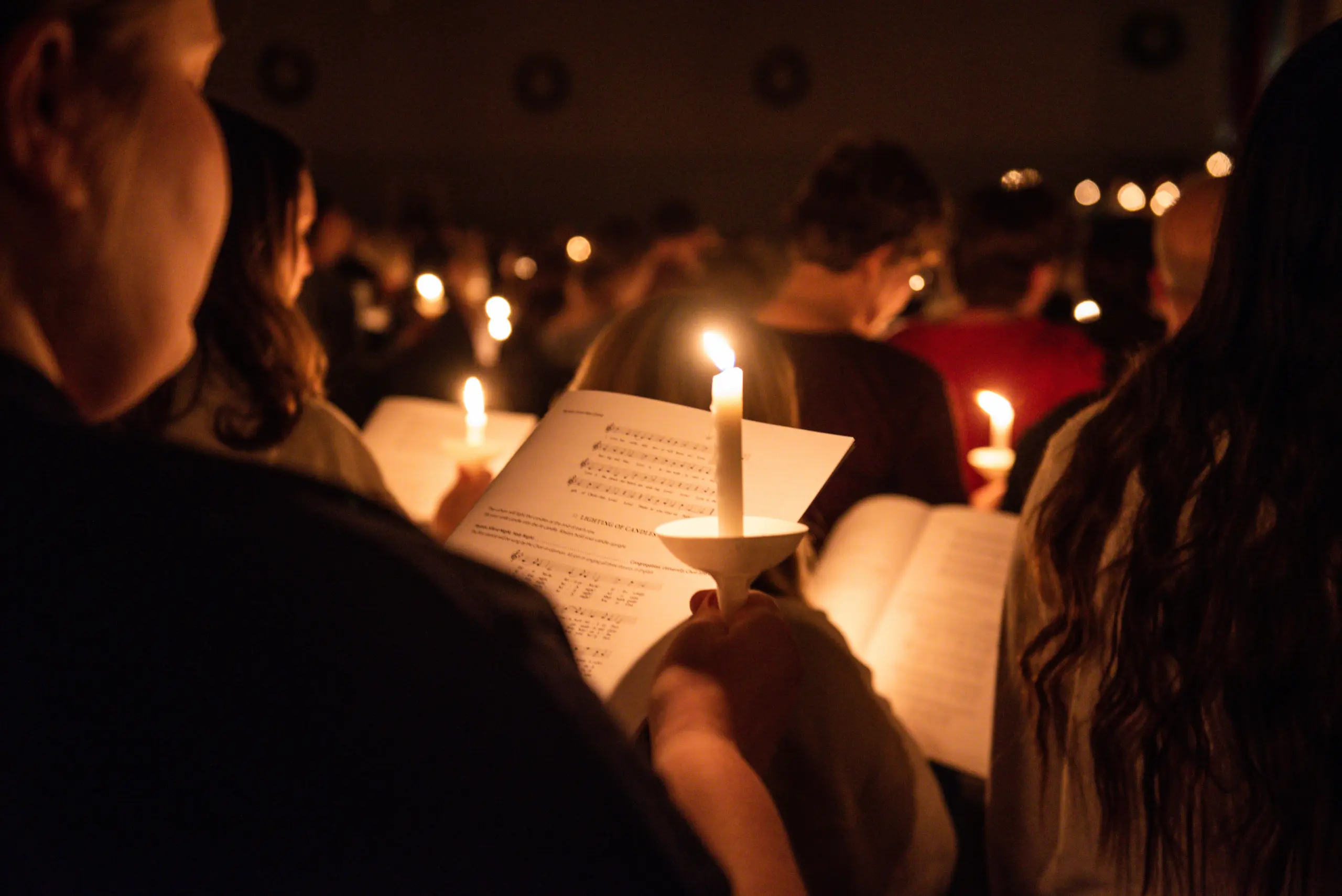 A group of people holding lit candles, reading from sheets of music, presumably during a nighttime or dimly lit vigil or musical event. The warm glow of the candles illuminates their hands and the sheets.