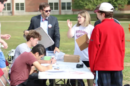 A group of people gathered around a table outdoors. One person in sunglasses and a suit holds a clipboard, while a young woman in a T-shirt gestures as she speaks. Several others are seated, writing on papers, with a few more standing around.
