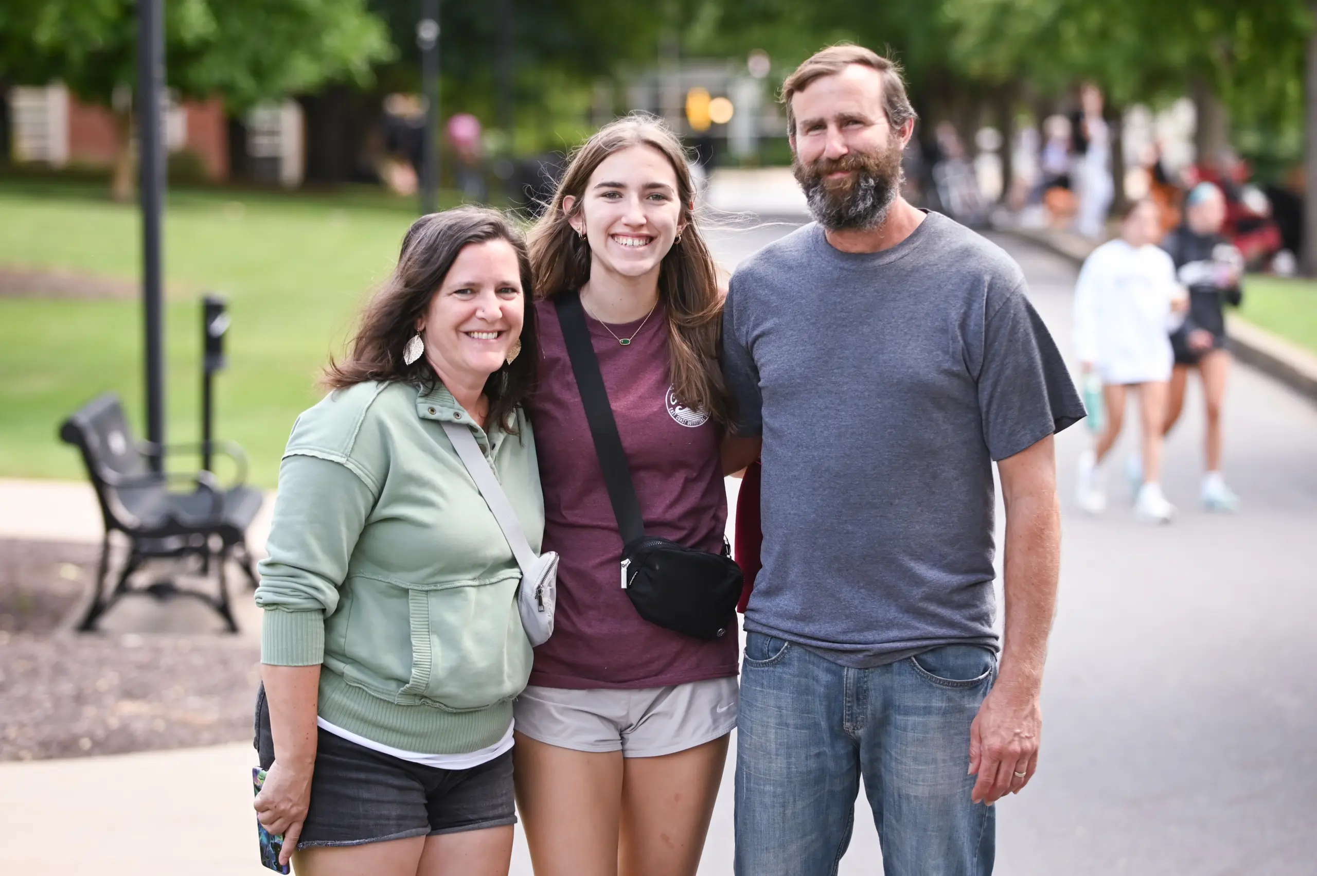 A group of three people stands outdoors on a path lined with trees. The person in the middle is wearing a maroon shirt and shorts, flanked by two adults in casual clothing. Blurred figures and benches are visible in the background.
