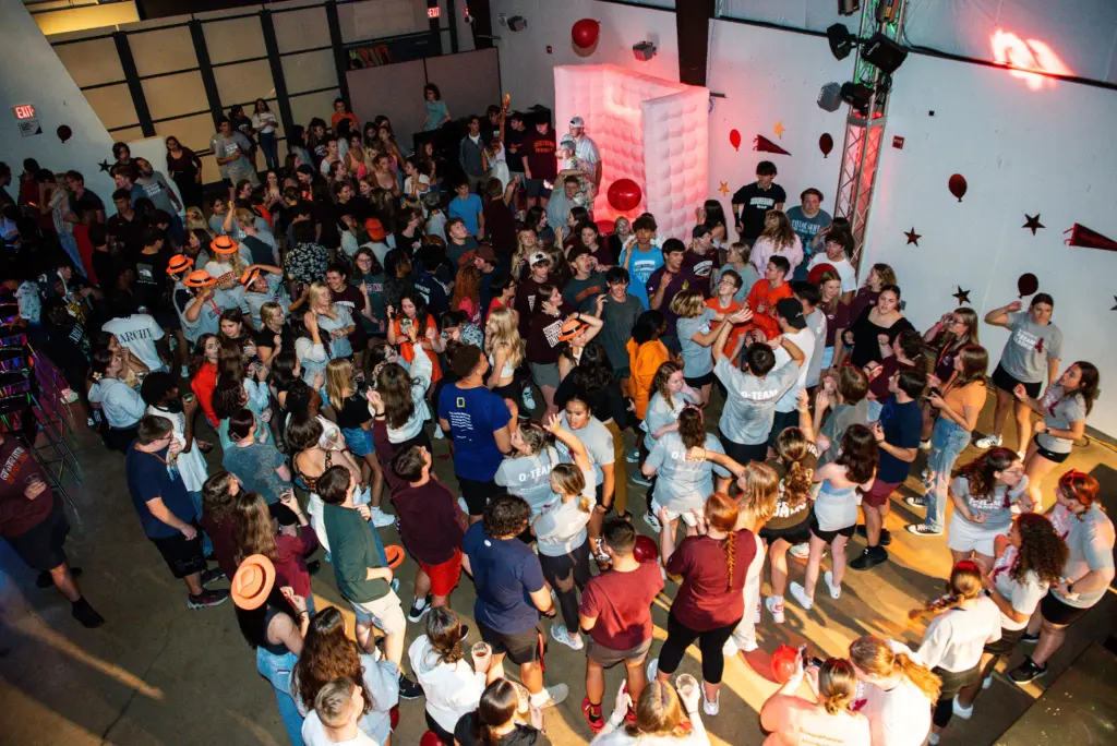A large crowd of people, mostly young adults, are gathered in a brightly lit indoor space. They are socializing and dancing. The walls are decorated with stars and balloons. The atmosphere is lively and festive.