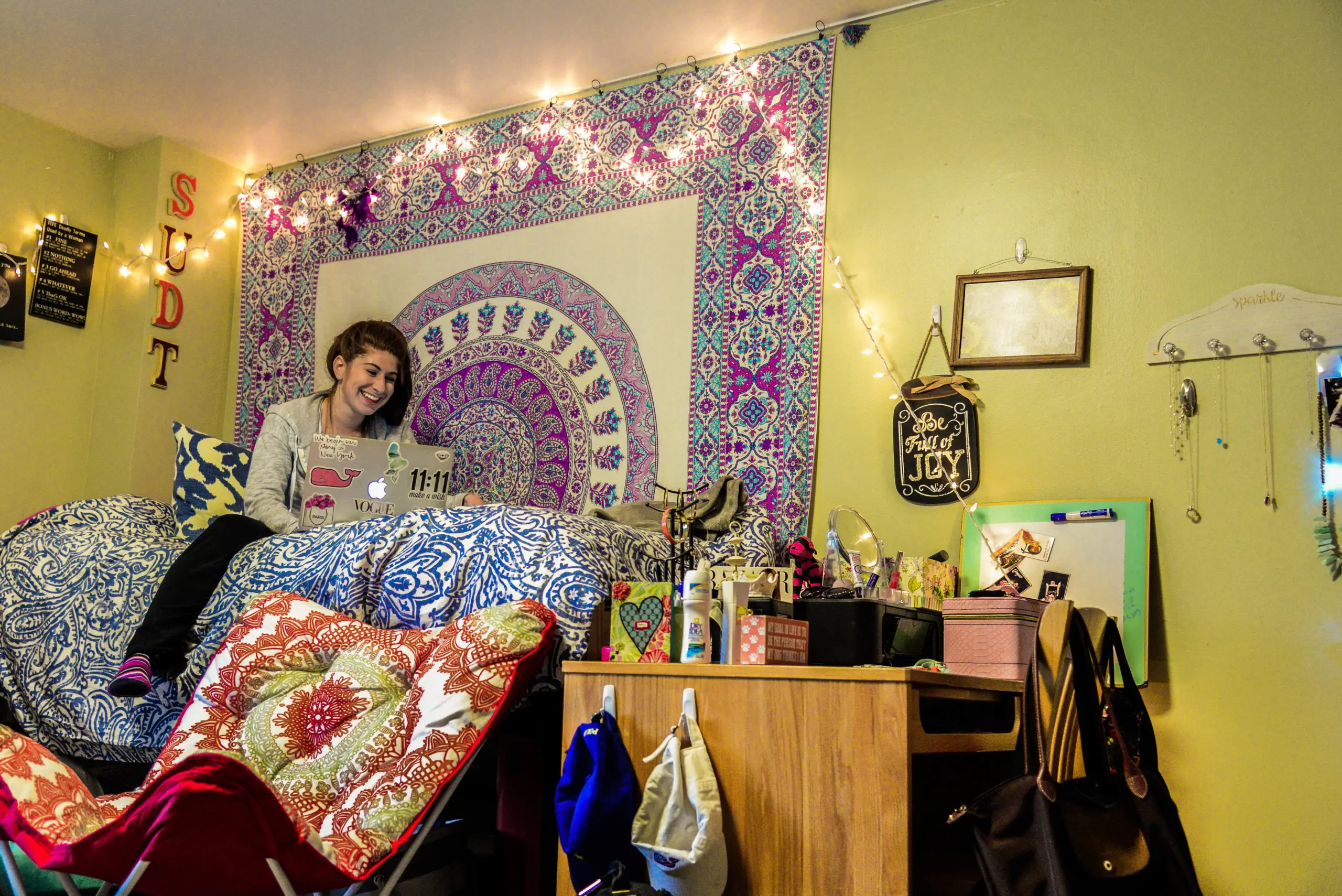 A cozy dorm room with a tapestry and string lights on the wall. A person sits on a bed with a laptop, surrounded by pillows. There's a small desk cluttered with books, snacks, and beauty products. .