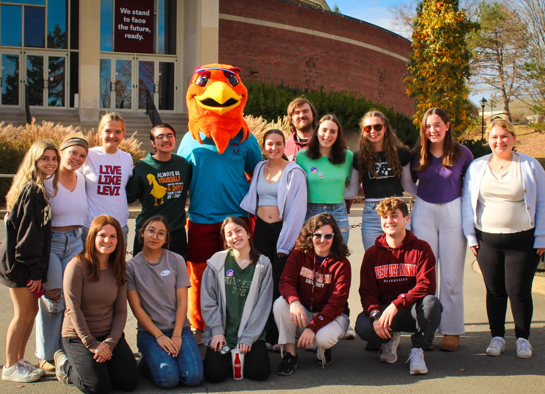 A group of 15 people pose outdoors with a colorful bird mascot. Some are sitting, while others stand. They are smiling and dressed in casual attire. A building and trees are visible in the background on a sunny day.