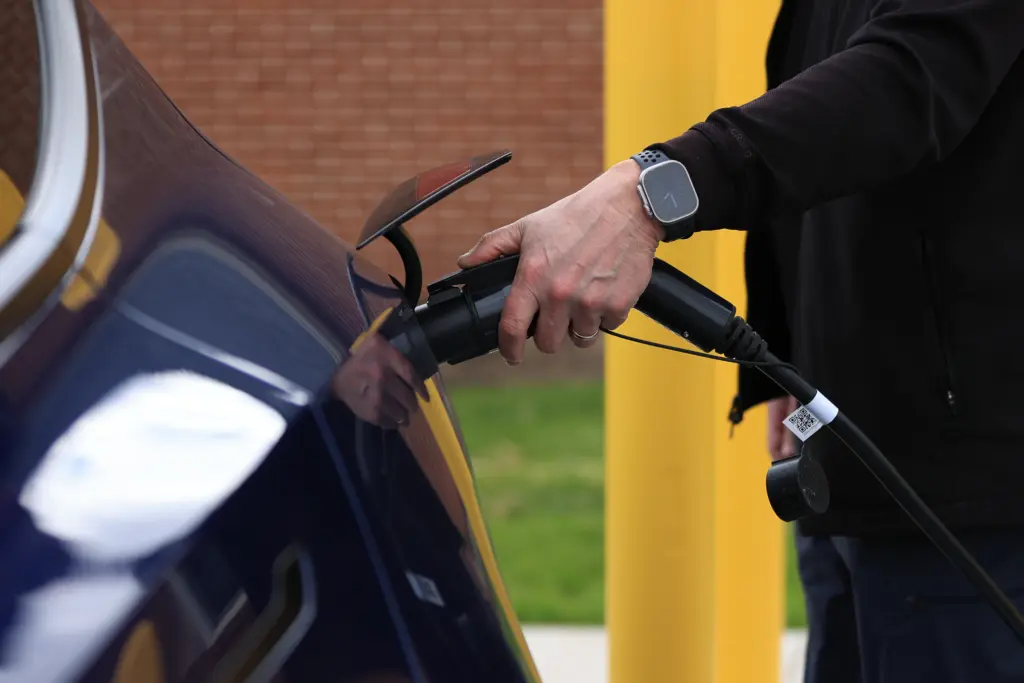 A person charges an electric car by plugging a charging cable into the vehicle. The car is blue, and the person wears a smartwatch on their wrist. The background shows a brick wall and a yellow post.