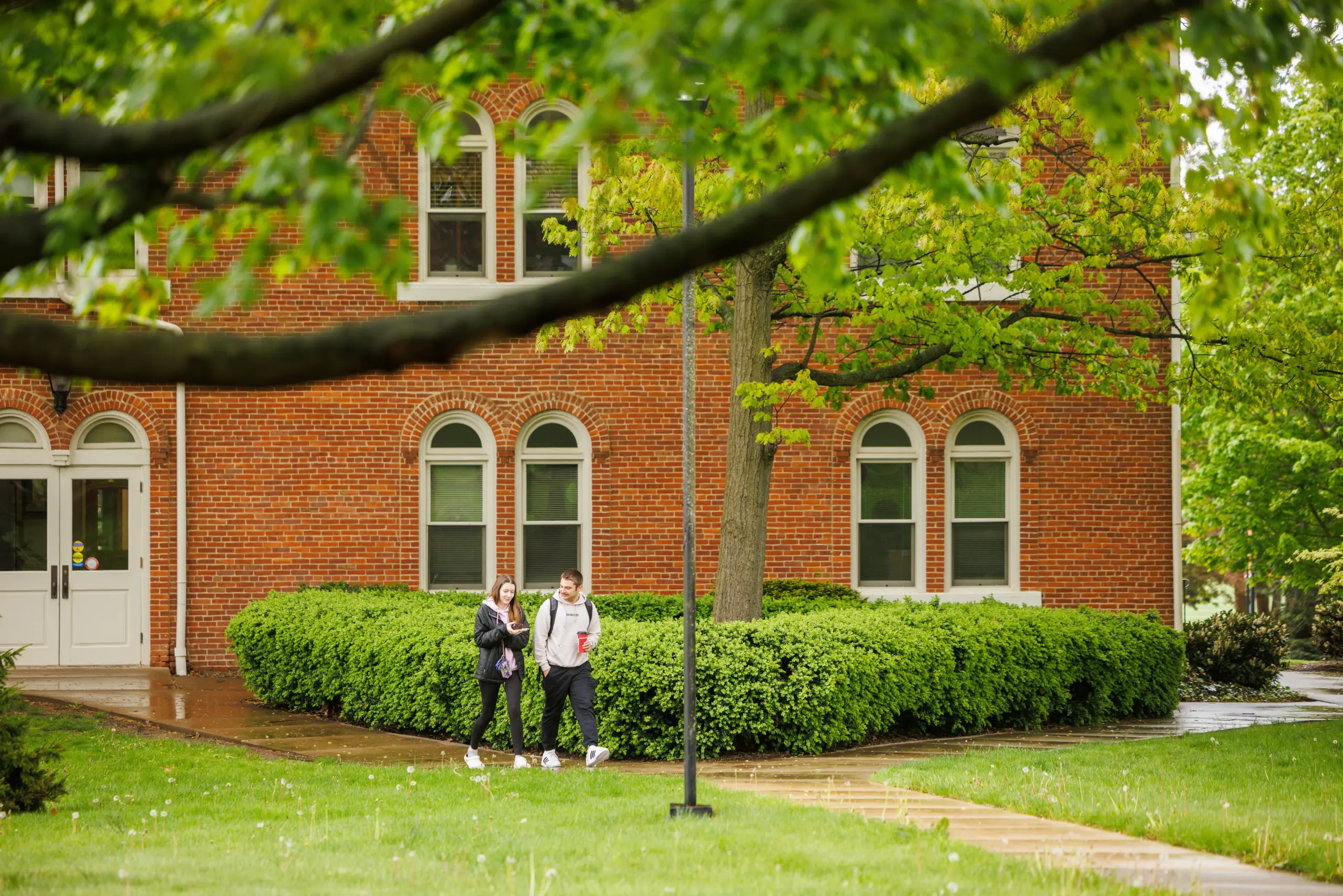 Two people walk on a pathway beside a red brick building with arched windows. The foreground has green grass and leafy branches, suggesting a park-like setting. The atmosphere appears calm and inviting.
