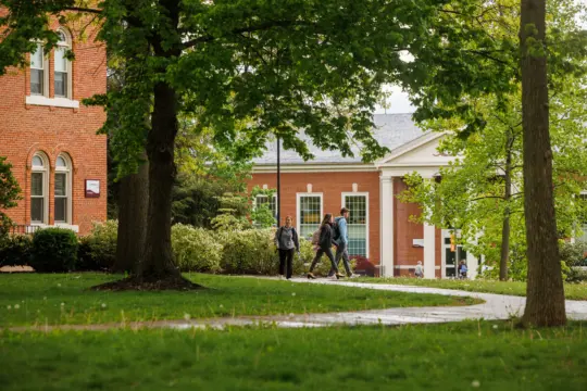 A group of people walk along a path in a college campus setting, surrounded by lush green trees and grass. Brick buildings with large windows are visible in the background, evoking a peaceful and academic atmosphere.